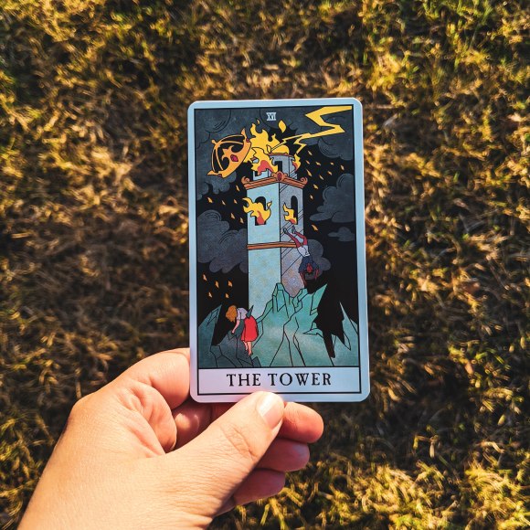Image of the author's hand holding a tarot card, "The Tower" from the deck "modern witch tarot" over a grassy background.