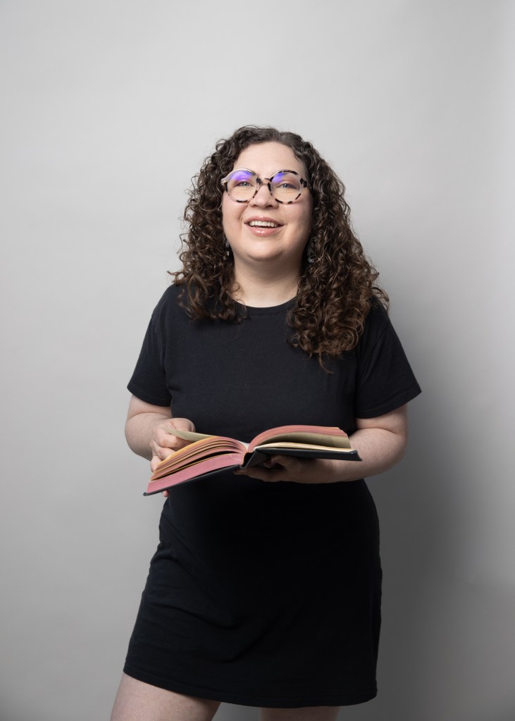Picture of Kaitlyn, a white woman with curly brown hair just past her shoulders. She wears glasses and a black shirt, and is smiling and laughing, a book held in her hands with red edged pages.