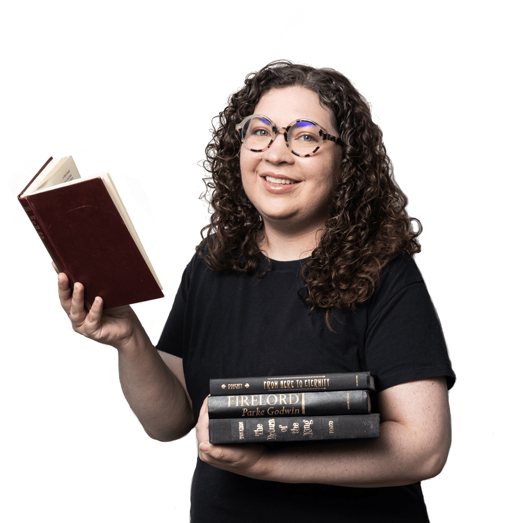 Picture of Kaitlyn, a white woman with curly brown hair just past her shoulders. She wears glasses and a black shirt, and smiles at the audience with a stack of books in her arms. Titles on the spines that are legible include "From here to eternity" and "Firelord" and "The Return of the King."